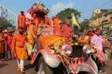 A vibrant parade features people dressed in colorful clothing, with many individuals in shades of orange, pink, and white, gathered around a vintage car adorned with flowers. The car is decorated with garlands and floral arrangements, while some participants have powdered colors on their clothes and skin, indicative of a festive celebration.
