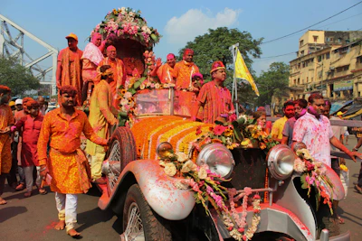 A vibrant parade features people dressed in colorful clothing, with many individuals in shades of orange, pink, and white, gathered around a vintage car adorned with flowers. The car is decorated with garlands and floral arrangements, while some participants have powdered colors on their clothes and skin, indicative of a festive celebration.