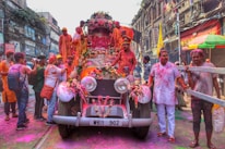 A group of people is celebrating a festival with vibrant colors, gathered around and on top of a vintage car adorned with flowers. The street is bustling with activity, and the participants are covered in colored powders, suggesting a festive mood. The surrounding buildings have an old architectural style, contributing to the lively and historic atmosphere.