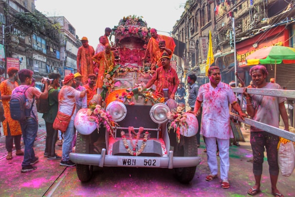 A group of people is celebrating a festival with vibrant colors, gathered around and on top of a vintage car adorned with flowers. The street is bustling with activity, and the participants are covered in colored powders, suggesting a festive mood. The surrounding buildings have an old architectural style, contributing to the lively and historic atmosphere.
