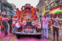 A group of people is celebrating a festival with vibrant colors, gathered around and on top of a vintage car adorned with flowers. The street is bustling with activity, and the participants are covered in colored powders, suggesting a festive mood. The surrounding buildings have an old architectural style, contributing to the lively and historic atmosphere.