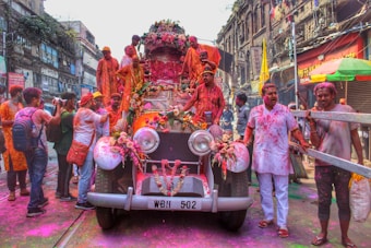 A group of people is celebrating a festival with vibrant colors, gathered around and on top of a vintage car adorned with flowers. The street is bustling with activity, and the participants are covered in colored powders, suggesting a festive mood. The surrounding buildings have an old architectural style, contributing to the lively and historic atmosphere.