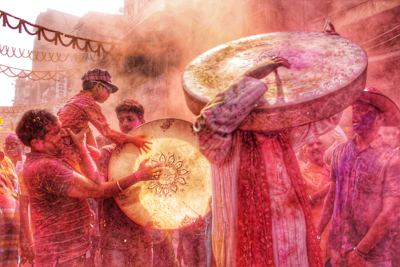 A vibrant scene filled with people celebrating with colored powders. Two large drums are prominently featured, with one person holding one over their head. The environment is festive, with people covered in colors and joyful expressions. Garlands are strung overhead, adding to the festive atmosphere.