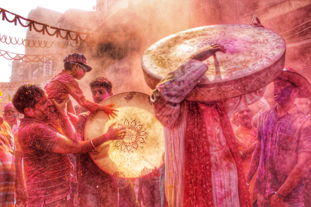 Traditional drums being played amidst a crowd celebrating Holi with blurred festive lights in the background.
