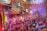 A crowded street filled with people celebrating a festival by throwing vibrant colored powders into the air. The buildings on either side are adorned with orange garlands, and there are numerous signs and wires overhead. Participants are enthusiastically engaging in the festivities, and there is a visible cloud of colors enveloping the scene.