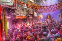 A crowded street filled with people celebrating a festival by throwing vibrant colored powders into the air. The buildings on either side are adorned with orange garlands, and there are numerous signs and wires overhead. Participants are enthusiastically engaging in the festivities, and there is a visible cloud of colors enveloping the scene.