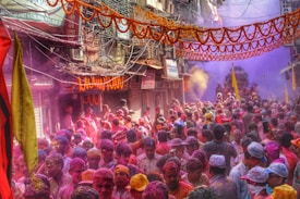 A crowded street filled with people celebrating a festival by throwing vibrant colored powders into the air. The buildings on either side are adorned with orange garlands, and there are numerous signs and wires overhead. Participants are enthusiastically engaging in the festivities, and there is a visible cloud of colors enveloping the scene.