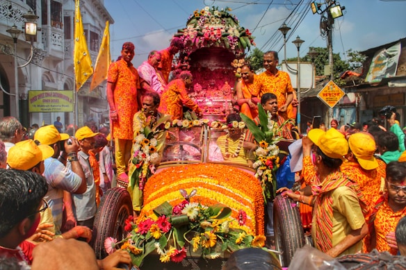 A vibrant parade with people dressed in colorful, ornate clothing surrounding a car lavishly decorated with flowers and garlands. The scene includes individuals wearing yellow caps, holding up flags, and a backdrop of buildings and signs. A joyful atmosphere is conveyed by participants interacting and taking photos.