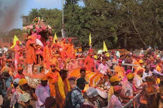 A lively and colorful festival parade with participants covered in bright powders, riding on a decorated float adorned with flowers. The crowd is vibrant, and the atmosphere is festive with flags and colorful garments.