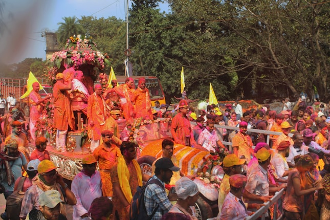 A lively and colorful festival parade with participants covered in bright powders, riding on a decorated float adorned with flowers. The crowd is vibrant, and the atmosphere is festive with flags and colorful garments.