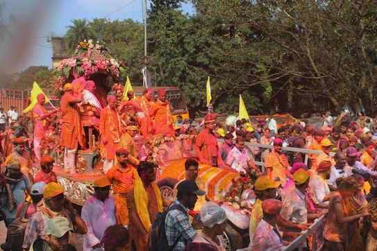 A lively and colorful festival parade with participants covered in bright powders, riding on a decorated float adorned with flowers. The crowd is vibrant, and the atmosphere is festive with flags and colorful garments.