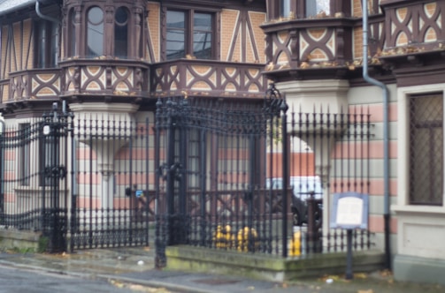 An ornate building facade featuring intricate brickwork and wooden designs, with large windows and decorative patterns. In front, there is a black wrought-iron fence with vertical bars, and a sign is visible near the entrance. The building displays a mix of reddish-brown and light beige hues, with a cobbled sidewalk beneath.