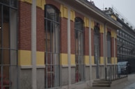 The image shows a building with an exterior made of red bricks, yellow panels, and large rectangular windows. There is a small set of stairs leading to a door, and scaffolding can be seen in the background, indicating construction or renovation work. A vehicle is parked near the stairs.