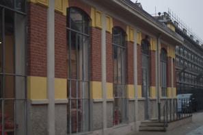 The image shows a building with an exterior made of red bricks, yellow panels, and large rectangular windows. There is a small set of stairs leading to a door, and scaffolding can be seen in the background, indicating construction or renovation work. A vehicle is parked near the stairs.