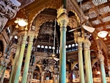 Intricately designed interior with ornate pillars and detailed carvings adorning the walls. A mix of gold and teal hues dominate the structural elements, with ceiling fans and vintage-style lights adding to the grandeur. Arched windows and elaborate wooden framing contribute to the opulent architectural style.