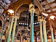 Intricately designed interior with ornate pillars and detailed carvings adorning the walls. A mix of gold and teal hues dominate the structural elements, with ceiling fans and vintage-style lights adding to the grandeur. Arched windows and elaborate wooden framing contribute to the opulent architectural style.