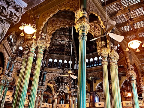 Intricately designed interior with ornate pillars and detailed carvings adorning the walls. A mix of gold and teal hues dominate the structural elements, with ceiling fans and vintage-style lights adding to the grandeur. Arched windows and elaborate wooden framing contribute to the opulent architectural style.