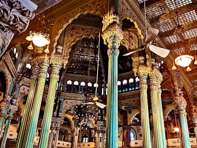 Intricately designed interior with ornate pillars and detailed carvings adorning the walls. A mix of gold and teal hues dominate the structural elements, with ceiling fans and vintage-style lights adding to the grandeur. Arched windows and elaborate wooden framing contribute to the opulent architectural style.
