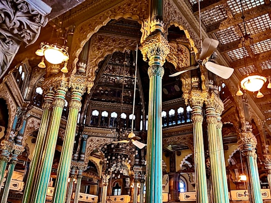Intricately designed interior with ornate pillars and detailed carvings adorning the walls. A mix of gold and teal hues dominate the structural elements, with ceiling fans and vintage-style lights adding to the grandeur. Arched windows and elaborate wooden framing contribute to the opulent architectural style.