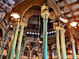 Intricately designed interior with ornate pillars and detailed carvings adorning the walls. A mix of gold and teal hues dominate the structural elements, with ceiling fans and vintage-style lights adding to the grandeur. Arched windows and elaborate wooden framing contribute to the opulent architectural style.