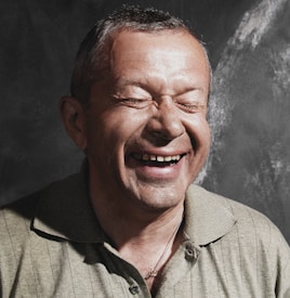 A man is laughing with his eyes closed, displaying a joyful expression. He is wearing a light-colored collared shirt, and the background is dark with a subtle textured pattern.