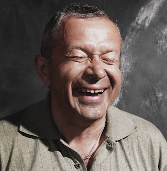 A man is laughing with his eyes closed, displaying a joyful expression. He is wearing a light-colored collared shirt, and the background is dark with a subtle textured pattern.