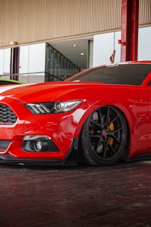 A red sports car with sleek, aerodynamic design is parked indoors. The car has tinted windows and black wheels with yellow brake calipers, reflecting a glossy finish under soft lighting. The surroundings include a modern structure with glass panels and red steel columns.