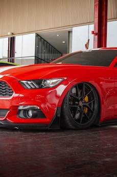 A red sports car with sleek, aerodynamic design is parked indoors. The car has tinted windows and black wheels with yellow brake calipers, reflecting a glossy finish under soft lighting. The surroundings include a modern structure with glass panels and red steel columns.