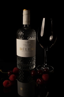 A glass wine bottle labeled 'Calabria Family Wines Bèlena True Partnership Rosé 2021' is placed alongside an empty wine glass. Surrounding the bottle are various berries, including cherries and raspberries, arranged on a reflective, dark surface which adds a dramatic ambiance to the image.