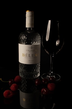 A glass wine bottle labeled 'Calabria Family Wines B&egrave;lena True Partnership Ros&eacute; 2021' is placed alongside an empty wine glass. Surrounding the bottle are various berries, including cherries and raspberries, arranged on a reflective, dark surface which adds a dramatic ambiance to the image.