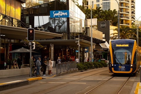 A modern urban street scene with a tram approaching a stop. Several people are waiting on the sidewalk, and there are shops and eateries around. The buildings feature large glass windows, and there are signs for various businesses. A traffic light is visible, and the street has clearly marked tram tracks.