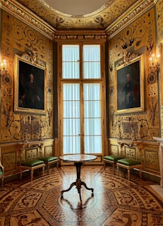 An opulent room adorned with intricate gold detailing on the walls and ceiling. Large portraits are framed on either side of a tall window with sheer curtains. The wooden floor features an elaborate inlaid pattern. Green upholstered benches line the walls, and a small round table stands in the center.