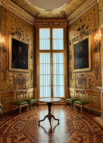 An opulent room adorned with intricate gold detailing on the walls and ceiling. Large portraits are framed on either side of a tall window with sheer curtains. The wooden floor features an elaborate inlaid pattern. Green upholstered benches line the walls, and a small round table stands in the center.