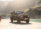 A rugged off-road vehicle is parked on a concrete dock by a body of water with mountainous terrain in the background. The vehicle is illuminated by soft sunlight, creating a warm and adventurous atmosphere.