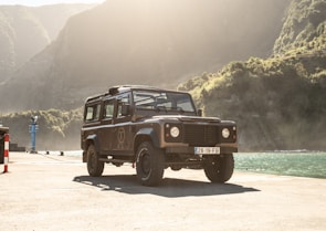 A rugged off-road vehicle is parked on a concrete dock by a body of water with mountainous terrain in the background. The vehicle is illuminated by soft sunlight, creating a warm and adventurous atmosphere.