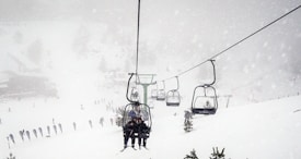 A ski lift is moving up a snowy mountain during a heavy snowfall. The sky is overcast, and there are several people visible on the lift and skiing on the slope below. Pine trees are partially covered with fresh snow.
