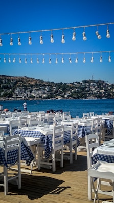 An outdoor restaurant setting featuring numerous white wooden tables and chairs, each covered with blue and white checkered tablecloths. Overhead, strings of light bulbs are strung across, with a backdrop of a sparkling blue sea and distant hillside buildings under a clear blue sky.