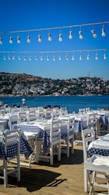 An outdoor restaurant setting featuring numerous white wooden tables and chairs, each covered with blue and white checkered tablecloths. Overhead, strings of light bulbs are strung across, with a backdrop of a sparkling blue sea and distant hillside buildings under a clear blue sky.