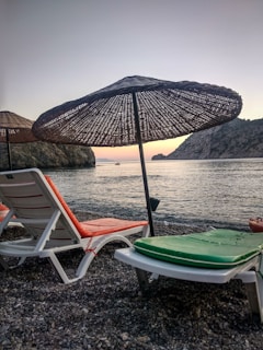 Two empty lounge chairs with colorful cushions are positioned on a pebble beach facing the calm sea. Large straw umbrellas provide shade, and the distant mountains create a serene backdrop. A small boat is visible on the water near the horizon, under a sky transitioning from light blue to hints of orange and pink at sunset.
