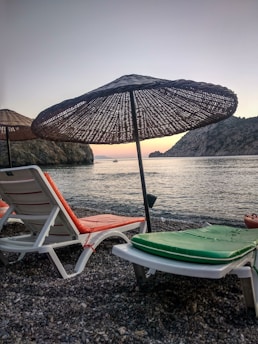 Two empty lounge chairs with colorful cushions are positioned on a pebble beach facing the calm sea. Large straw umbrellas provide shade, and the distant mountains create a serene backdrop. A small boat is visible on the water near the horizon, under a sky transitioning from light blue to hints of orange and pink at sunset.