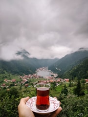 A scenic view of a lush green valley with a lake surrounded by mountains and a village. In the foreground, a hand holds a traditional Turkish tea glass on a saucer, focusing on a cultural tea moment amidst nature.
