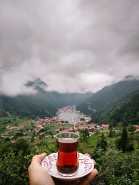 A scenic view of a lush green valley with a lake surrounded by mountains and a village. In the foreground, a hand holds a traditional Turkish tea glass on a saucer, focusing on a cultural tea moment amidst nature.