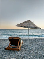 A single wooden lounge chair with a cushion sits on a pebble beach, facing a calm sea. A thatched umbrella provides shade under a clear sky with a gentle gradient from deep blue to orange near the horizon.