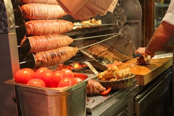 A street food setup featuring a large rotisserie with layers of meat being cooked on skewers. Fresh red tomatoes are in a metal container, while a basket holds pieces of bread. A person is slicing cooked meat on a wooden cutting board.