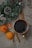 A warm and cozy setting with a cup of dark beverage placed on a sheet of old newspaper. On the side, there are sprigs of evergreen with pinecones and small red and white berries, evoking a winter or holiday theme. Fresh mandarins and cinnamon sticks are also laid out, adding a rustic and aromatic touch.