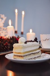 A slice of layered cake with white frosting and chocolate shavings on top sits elegantly on a white plate. In the background, there are lit candles of varying heights creating a warm, cozy ambiance. To the side, a cup appears partially visible, and decorative elements like pine cones and red berries add a festive touch.