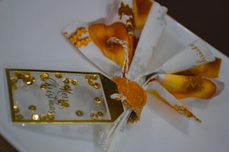 A festive, decorative item featuring a fabric napkin elaborately folded and tied with a gold and white string. It includes a tag with the words 'Merry Christmas' surrounded by gold sequins, and a golden wax seal, all placed on a white plate.