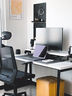 A modern workspace featuring a sleek black office chair, a large monitor on a stand, and a laptop on a desk with a white top and black legs. The wall is adorned with a minimalist artwork and shelving with various items. A mustard-colored ottoman is positioned beneath the desk, and a small potted plant is visible on the windowsill.