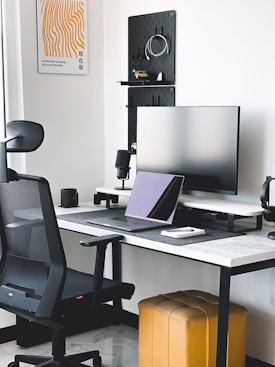 A modern workspace featuring a sleek black office chair, a large monitor on a stand, and a laptop on a desk with a white top and black legs. The wall is adorned with a minimalist artwork and shelving with various items. A mustard-colored ottoman is positioned beneath the desk, and a small potted plant is visible on the windowsill.