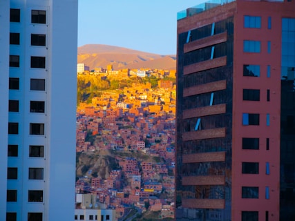 A densely built residential area with numerous small houses is nestled between two modern skyscrapers. The buildings in the foreground are tall structures with reflective surfaces, while the background features a hillside bathed in warm, golden light, creating a striking contrast between urban architecture and traditional housing.
